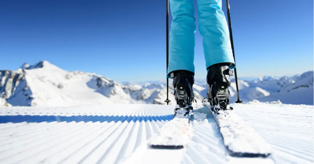 A close-up of a woman wearing light blue snow pants and downhill skis on a freshly groomed ski slope