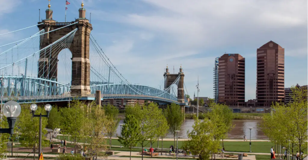 A large bridge made of stone, with suspensions painted bright blue, spanning the Ohio River, with the Ohio River Trail in the foreground and two large copper-colored buildings in the background.