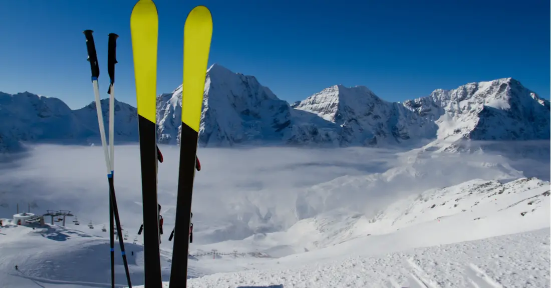 A pair of skis and ski poles propped up with snow-covered mountains in the background.