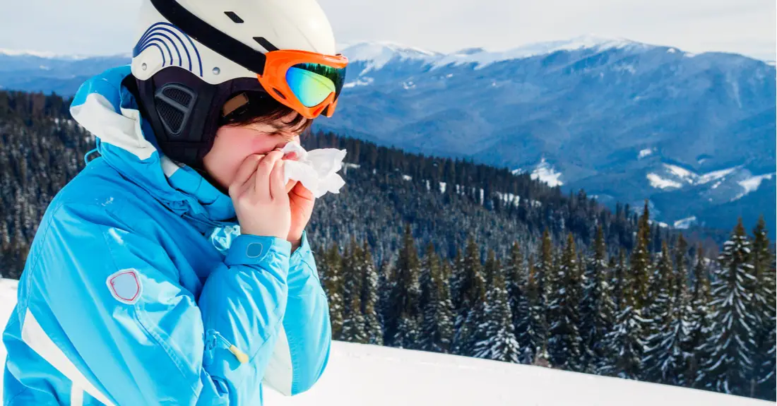 A woman in a ski jacket, ski helmet and ski goggles blowing her nose with a tissue she packed in her pocket.