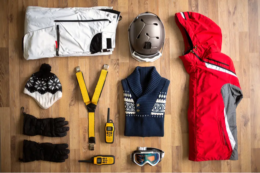 Ski clothes and gear, including a jacket, a helmet, snow pants, ski gloves and ski goggles, folded on a wooden floor, ready to be packed.